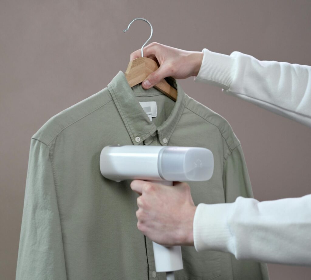 Our Amazon Travel Essentials Hands steaming a green shirt with a portable steamer against a grey background.